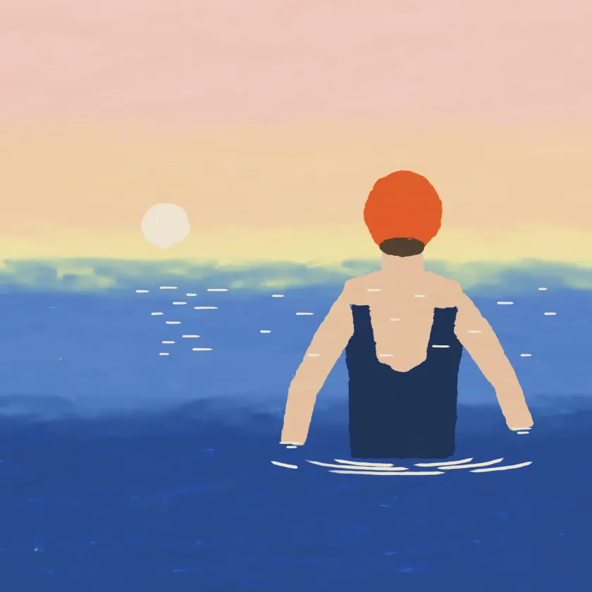 A woman in an orange swim cap wading into a cold blue sea at sunrise.